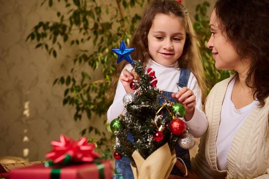Mother and daughter decorating an eco Christmas tree with beautiful shiny bauble and Xmas toys. Christmastime. New Year's preparations and celebration. Enjoying happy winter holidays in family at home