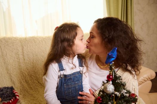 Happy multi-ethnic family of loving mother and daughter kissing, hugging together while enjoying Christmas preparations at home, decorating Christmas tree. Magic festive atmosphere of winter holidays