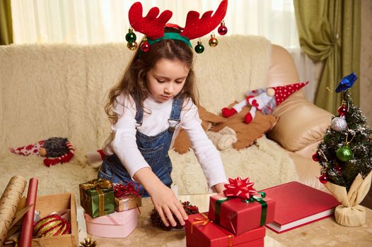 Adorable Caucasian little girl wearing deer antler hoop and denim overalls, standing at a table with Christmas gifts, wrapping materials for packing presents. Xmas. Happy New Year and winter holidays