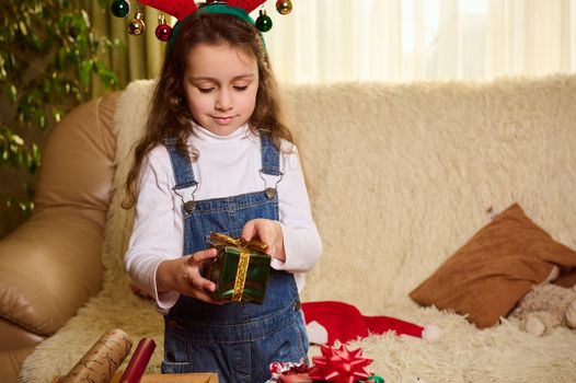 Adorable child, cute baby girl with deer antler hoop on her head, holds a beautiful gift box, enjoying the New Year's preparations at home. Happy festive atmosphere of the upcoming winter holidays