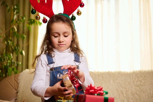 Caucasian child, adorable baby girl with deer antler hoop on her head, holding a Christmas gift. Family, happy winter holidays and beautiful people concept. New Year. Christmas Time. December 25th