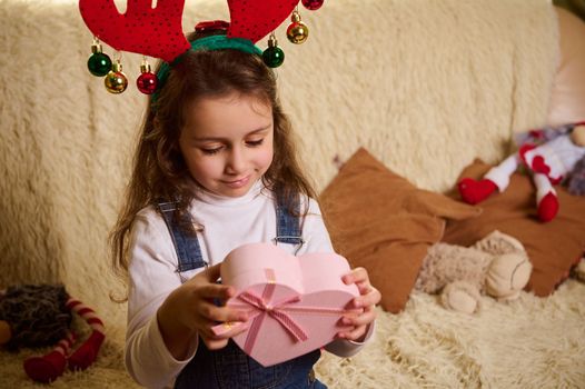 Charming Caucasian little child girl wearing a deer antler hoop, holding a pink heart shaped gift box. Boxing Day. Happy winter holidays. Happy New Year. Merry Christmas. Christmastime