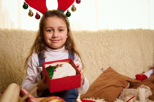 Close-up portrait of a Caucasian charming little child, adorable baby girl smiling a beautiful toothy smile while unpacking a cute Christmas present. Happy winter holiday. New Year's celebration