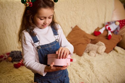 Adorable little child girl wearing deer antler hoop, unpacking a beautiful pink heart shaped gift box. Happy magic atmosphere of winter holidays. New Year's event. Merry Christmas. Christmastime