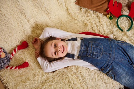 Top view lovely child girl in blue denim overalls, cutely smiles looking at camera, lying on a comfortable sofa at home, surrounded by Christmas items. Happy atmosphere of the upcoming winter events