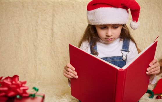 Portrait of a cute little child girl in Santa's hat, with a hardcover red book, reading fairy tales at cozy home interior with Christmas decors. Copy advertising space for promotional text. New Years