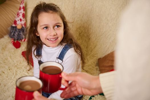 Adorable Caucasian little smiling a cheerful toothy smile looking at her mom bringing her hot chocolate drink. Food and drink. Festive sweet beverage. Happy winter holidays. Christmas Time