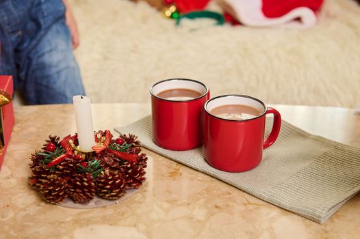 Red cups with hot cocoa drink on a green napkin, next to a decorative Christmas lit candle on marble table. New Year atmosphere at home.