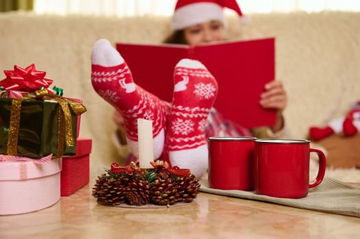 Selective focus on red socks with Christmas pattern, on blurred background of a pretty woman in Santa's hat reading book, relaxing on a comfortable sofa during New Years winter holidays. Copy ad space