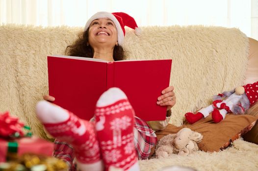 Attractive happy woman in Santa hat and socks with Christmas pattern, reading fairy tales, sitting on sofa at home with blurred gifts in foreground. Youth in the heart. Magical mood of winter holidays