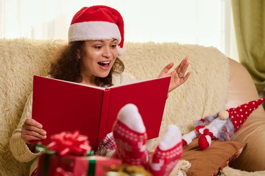 Beautiful delightful Hispanic woman in Santa hat and socks with Christmas pattern, reading fairy tales at home, with blurred presents in foreground. Magical festive atmosphere during winter holidays