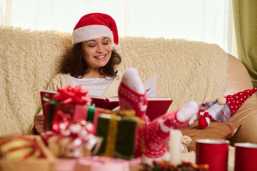 Dark-haired multi-ethnic delighted pretty woman in Santa's hat, reads fairy tales while rests at home during winter holidays. Remembering happy moments from childhood. Youth at heart. Christmas mood