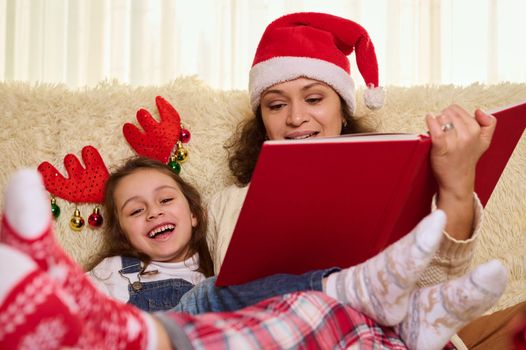 Happy multi-ethnic family, a loving mother in Santa's hat and lovely daughter wearing deer antler hoop, enjoying happy moments together by reading fairy tales, during Christmas holidays at home