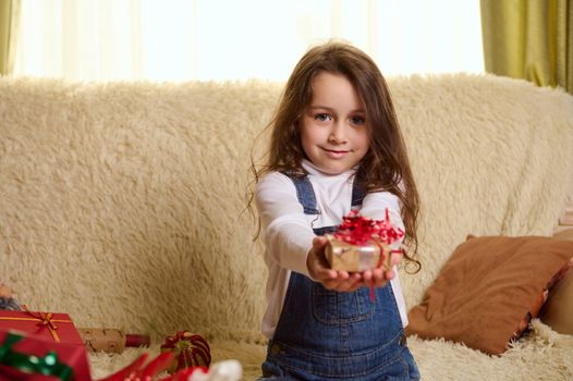 Portrait of beautiful European little girl dressed in white pull over and denim overalls, cutely smiles while handing to camera a Christmas gift, sitting on a comfortable sofa in a cozy home interior.