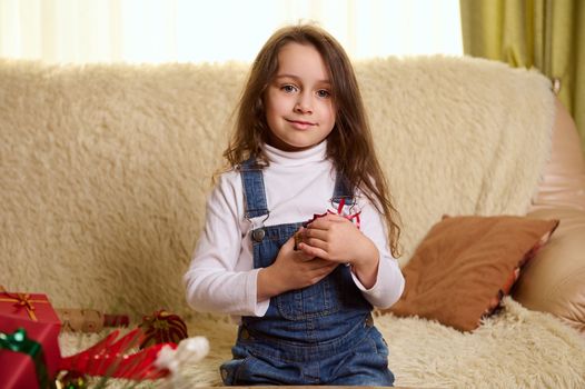 Adorable Caucasian child, a little girl wearing a white pull over and denim overalls, cutely smiles to camera, gently hugging her Christmas gift, sitting on a comfortable couch in a cozy living room