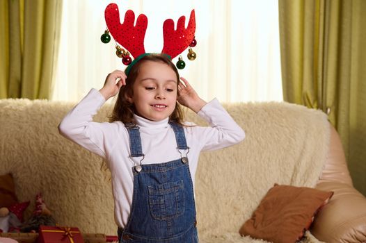 Adorable child, a Caucasian little girl in denim overalls and white pull, puts on antler deer hoop, enjoys Christmas atmosphere at home. Happy winter holidays. Magic mood. December 25. Happy childhood