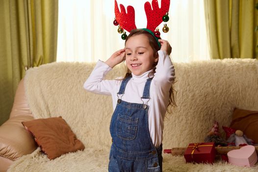 Cute happy child, Caucasian little girl having fun at cozy home interior, during Christmas holidays, putting on deer antler hoop, smiling a beautiful cheerful toothy smile, enjoying festive atmosphere