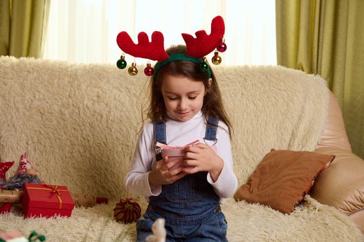 Beautiful Caucasian little child girl in blue casual denim overalls and funny deer antler hoop, holding a cute Christmas present in her hands, sitting on a comfortable sofa in the living room