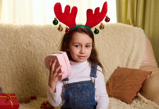 Beautiful Caucasian little girl wearing funny deer antler hoop, holding a heart shaped pink gift box, sitting on a comfortable in living room, enjoying the upcoming winter holidays. Merry Christmas