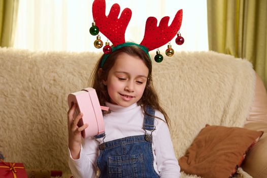 Charming cutie, adorable European little girl in deer antlers hoop, shakes a pink heart shaped gift box, trying to guess its contents. Happy surprises and presents for Christmas or New Year