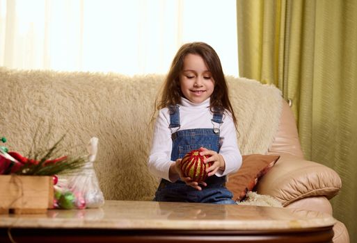 Adorable Caucasian little child girl in blue denim overalls, sitting on a comfortable sofa in the living room, surrounded by festive decorations for upcoming winter holidays and Christmas. Magic mood