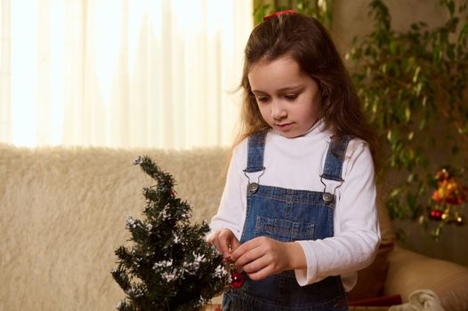 Close-up portrait of a Caucasian pretty child, a serene little girl with long hair, decorates a small Christmas tree with shiny balls, enjoys upcoming winter holidays. Xmas and New Years preparations