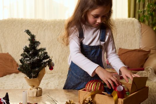 Delightful little girl in white shirt and blue denim overalls, standing at a journal marble table with Christmas toys in wooden crate, enjoying New Year and Xmas preparations in a cozy home interior