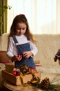 Adorable little child girl standing by journal table with toys in wooden box, chooses balls for decorating Christmas tree, prepares for New Year's event during winter holidays, in cozy home interior
