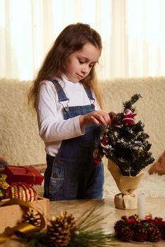 Adorable European little girl in blue denim overalls, stands by a journal table in the cozy living room, enjoys New years preparations, decorates a small Christmas tree with toys. Winter holidays