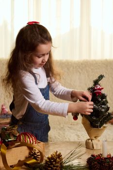 Adorable Caucasian little child girl with long hair, dressed in white pull and blue denim overalls, decorates a small eco Christmas tree with beautiful toys. New Year's preparations. Winter holidays