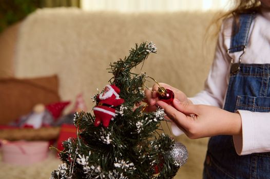 Close-up kid's hand with a Christmas toy, a shiny ball while decorates a Christmas tree. December 25. New Years preparations. Enjoying happy winter holidays. Magic festive atmosphere at home