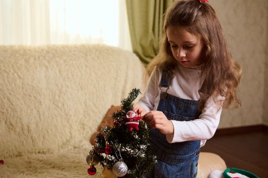 Adorable Caucasian little girl holding a shiny ball while hanging decorative toys on a Christmas tree in the cozy home interior. New Year's preparations. Magic festive atmosphere of winter holidays