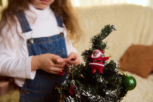 Selective focus on a small eco-Christmas tree, against a blurred background of cute child girl hanging decorative toys and balls while decorates it for the winter holidays. New Year's preparations