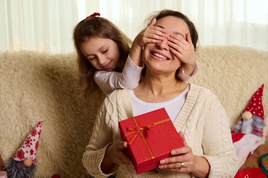 Pretty loving mother smiling a cheerful toothy smile, while her lovely daughter covers her eyes with her hands, offers her a Christmas present in stylish red box, sitting together on sofa at home