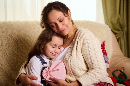 Portrait of a happy multiethnic woman, a caring loving mother, tenderly hugging her beautiful lovely daughter, holding a heart-shaped pink gift box. Little girl enjoys spending time with mom at home
