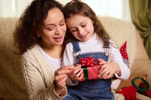 Close up portrait of beautiful loving mother - multi-ethnic woman, tenderly hugging her adorable little daughter unpacking Christmas present. Winter New Year holidays. Happy childhood and motherhood