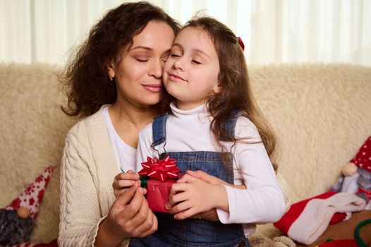 Moments of tenderness. Gorgeous dark-haired multi-ethnic pretty woman, a caring loving mom hugs her little daughter, while exchanging Christmas presents at home. Childhood. Happy family relationships