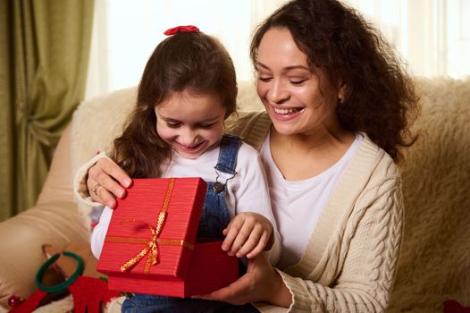 Happy little girl, lovely daughter sitting on her mom's knees and experiencing happiness, joy and excitement while opens the red gift box with Christmas present. Magic atmosphere at winter holidays