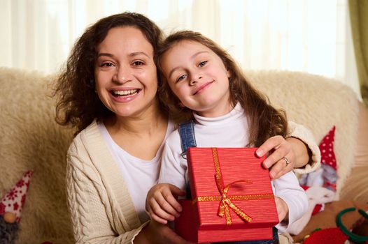 Portrait of a delightful caring mother and her cute little daughter embracing, smiling cheerful toothy smile looking at camera while opening Christmas gift. Happy loving family. Carefree childhood