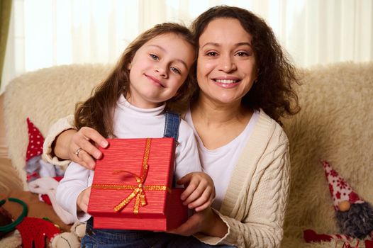 Happy multi-ethnic millennial family, loving mother and her adorable little daughter smiling a cheerful smile, looking at camera while unpacking Christmas gifts, sitting on sofa in cozy home interior