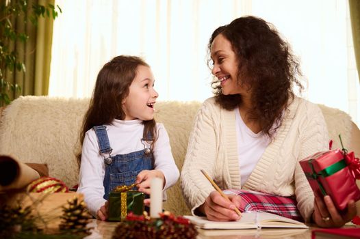 Adorable mother and daughter make a gift list, wrapping Christmas presents, sitting on couch surrounded by Xmas decorations. Preparing for the New Year at home. Festive mood during winter holidays