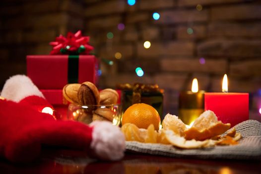 Close-up Still life. Christmas food background with ripe juicy fresh tangerines on festive table with beautiful presents, cookies and lit decorative red and green candles, against sparkling garland