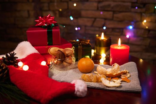 Magical atmospheric Christmas background with gifts, lit candle, peeled fresh juicy tangerines against brick wall, with hanging sparkling colorful lights. Happy New Year. Merry Xmas. Winter holidays