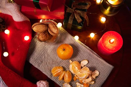 Top view of peeled tangerines on a green napkin, next to homemade delicious pastries, gifts and sparkling garland lying on a red santa hat, candlelight. Christmas still life. New Year's preparations