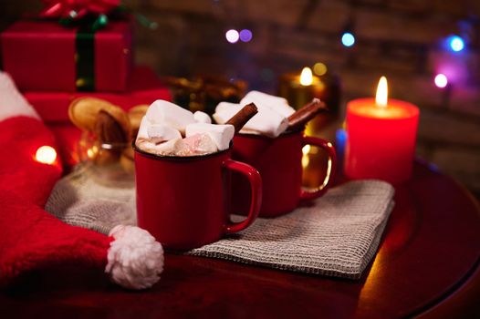 Food still life. Red cups of hot chocolate cocoa drink with marshmallows and cinnamon, on festive Christmas table against blurred background of lit candles, Xmas presents and sparkling lights garlands
