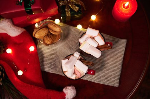 Food still life. Mugs of hot chocolate drink with marshmallows and cinnamon, on festive Christmas table against blurred background of lit candles, Xmas gifts and bright lights on garlands. Top view