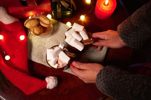 Overhead view of woman's hands, putting hot cocoa drink with marshmallows on the green napkin on a wooden festive Christmas table, with delicious homemade cookies and garland lights, in candlelight