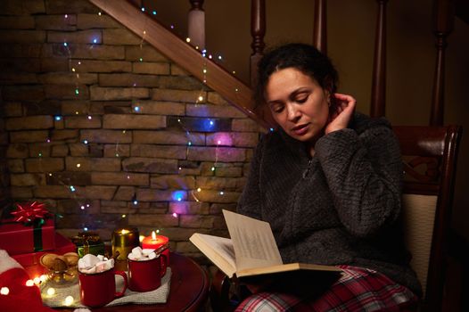 Relaxed woman reads book in candlelight, sitting by table with cups of hot cocoa drink with marshmallows, Christmas gifts, beautiful Xmas lights on Santa's hat and sparkling garlands on a brick wall.