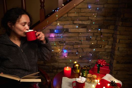 Close-up portrait of charming multi-ethnic woman drinking hot chocolate or cocoa with marshmallows, sitting by table in cozy home interior for Christmas. Winter holiday. December 25th. Happy New Year