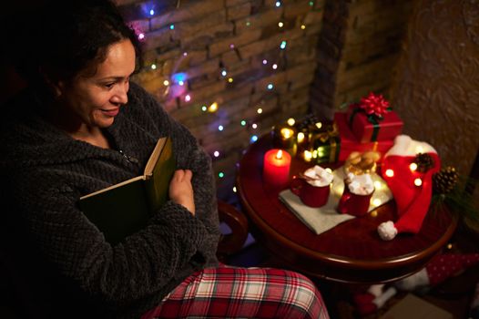 Top view of delightful charming woman with book, enjoying reading by candlelight, sitting at table with Christmas gifts, cups of hot cocoa with marshmallows and sparkling garlands. Merry Christmas.